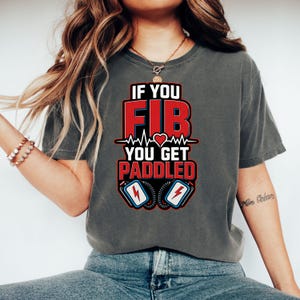 Könnte beinhalten: Dunkelgraues T-Shirt mit dem Text "IF YOU FIB YOU GET PADDLED" in Rot und Weiß. Das Design umfasst eine Herzschlaglinie, ein Herz und zwei Defibrillator-Paddles mit Blitzen. Ein medizinisches Grafik-T-Shirt.