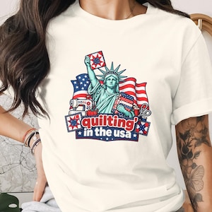May include: A cream-coloured t-shirt featuring a graphic of the Statue of Liberty holding a quilt block, with a sewing machine, scissors, and an American flag in the background. The text "quilting in the USA" is printed in red and white letters.