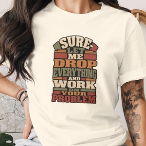 Puede incluir: Una camiseta de manga corta de color crema con un estampado gráfico multicolor que incluye el texto: SURE LET ME DROP EVERYTHING AND WORK ON YOUR PROBLEM. Las letras están dispuestas en bloques apilados en tonos rojo, naranja y verde oliva.