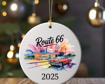 Op de afbeelding: Een witte keramische kerstbal met een Route 66 diner-scène en een rode cabriolet. De bal heeft de tekst "Route 66" en "2025" in het zwart. De achtergrond heeft kleurrijke accenten in aquarel-stijl.