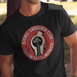 May include: Black t-shirt with a circular design. The design features the text "BIONIC SHOULDER CLUB CUSTOM PARTS INSTALLED" around an illustration of a bionic shoulder joint in red, beige, and black.