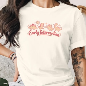 May include: A cream-colored short-sleeve t-shirt featuring a graphic print of four small dinosaurs, a sun, and stars in shades of pink and orange. Below the dinosaurs, the text "Early Intervention" is printed in a cursive pink font.