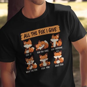 May include: A black short-sleeved t-shirt featuring a graphic of six cartoon foxes with the text "ALL THE FOX I GIVE:" at the top. Below the foxes are the phrases "FOX IT", "FOR FOX SAKE", "FOX EVERYONE", "WHAT THE FOX", "FOX YOU", and "HOLY FOX".
