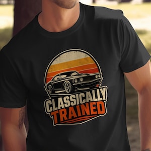 Może przedstawiać: Czarny t-shirt z krótkim rękawem z grafiką klasycznego samochodu sportowego na tle koła w kolorach zachodu słońca. Pod samochodem znajduje się napis "CLASSICALLY TRAINED" wydrukowany pogrubionymi literami w kolorze pomarańczowym i białym.