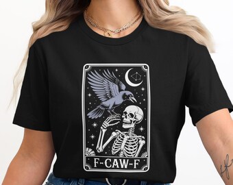 May include: Black t-shirt with a graphic design of a raven perched on a skeleton's hand, set against a starry night sky with a crescent moon. The text "F-CAW-F" is at the bottom. The design is in a rectangular frame.