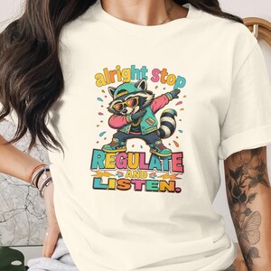 May include: A cream-coloured t-shirt featuring a graphic of a raccoon wearing a cap, sunglasses, and a jacket, performing a dabbing pose. The text reads "alright stop REGULATE AND LISTEN." The design uses vibrant colours including teal, pink, and yellow.