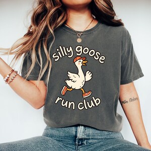 May include: A charcoal gray t-shirt featuring a graphic of a white goose wearing an orange baseball cap and orange sneakers. The text above the goose reads silly goose and the text below reads run club in a rounded, cream-colored font.