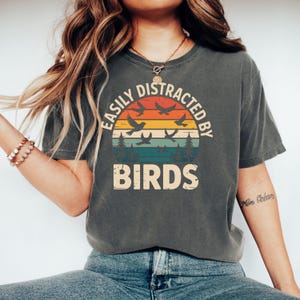 Könnte beinhalten: Dunkelgraues T-Shirt mit dem Text "EASILY DISTRACTED BY BIRDS" in einem kreisförmigen Design. Das Design zeigt einen Sonnenuntergang mit fliegenden Vögeln und Bäumen. Das Shirt ist ein lässiger, bequemer Stil.
