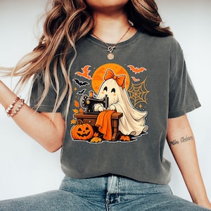 Könnte beinhalten: Ein anthrazitfarbenes T-Shirt mit einer Grafik eines weißen Geistes mit orangefarbener Schleife, der an einer Nähmaschine im Vintage-Stil näht. Das Design zeigt einen orangefarbenen Vollmond, schwarze Fledermäuse, ein Spinnennetz und einen Kürbis.