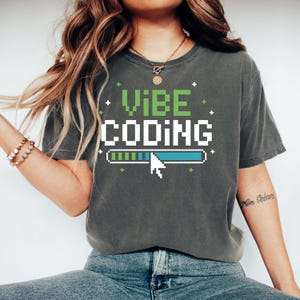 Puede incluir: Camiseta gris oscuro con las palabras "VIBE CODING" en fuente pixelada verde y blanca. Un gráfico de barra de carga con un cursor está debajo del texto. La camiseta es de estilo casual.
