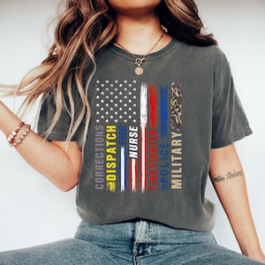 May include: A charcoal gray t-shirt featuring a graphic of the American flag composed of vertical stripes. The design includes the text CORRECTIONS, DISPATCH, EMS, NURSE, FIREFIGHTER, POLICE, and MILITARY in various colors alongside a camouflage pattern.