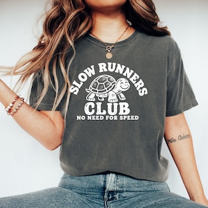 May include: A charcoal gray short-sleeve t-shirt featuring a white graphic of a turtle. The text reads SLOW RUNNERS CLUB and NO NEED FOR SPEED. The design is centered on the chest of the garment, which has a crew neckline and a relaxed fit.