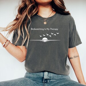 May include: Dark gray t-shirt with the text "Birdwatching Is My Therapy" above a graphic of a sunset and flying birds. The shirt is a comfortable, casual style.