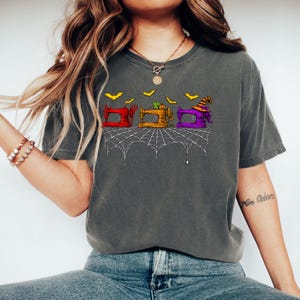 Könnte beinhalten: Ein anthrazitfarbenes T-Shirt mit einem Grafikdruck von drei Nähmaschinen in Rot, Orange und Lila. Die Maschinen sind mit einem Kürbis und einem Hexenhut verziert, umgeben von gelben Fledermäusen und einem weißen Spinnennetz-Design am unteren Rand.