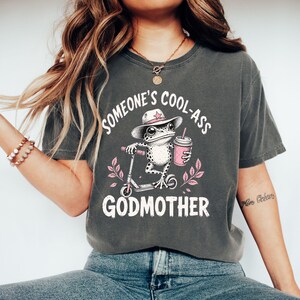 Op de afbeelding: Een antracietgrijs t-shirt met een afbeelding van een kikker met een roze cowboyhoed op een step, die een roze drankje vasthoudt. De tekst SOMEONE'S COOL-ASS GODMOTHER staat in witte letters, omringd door roze bladillustraties.