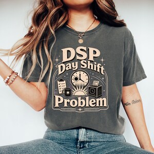 Może przedstawiać: Ciemnoszary t-shirt z krótkim rękawem z nadrukiem graficznym z napisem "DSP Day Shift Problem". Wzór zawiera zegar, podkładkę z klipsem, kubek do kawy, ikonę słońca i zegar rejestrujący czas, wszystko w kolorze kremowym i czarnym.