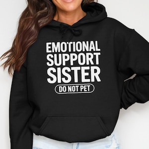 May include: Black hoodie with the white text "EMOTIONAL SUPPORT SISTER DO NOT PET". The sweatshirt features a front pocket and a drawstring hood. The text is centered on the chest.