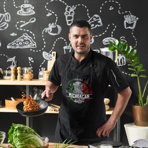 May include: A man wearing a black apron with a Mexican flag design and the text "Estados Unidos Mexicanos Michoacan". He is holding a black frying pan with food in it.