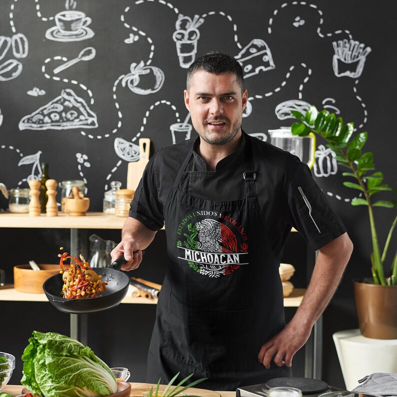 May include: A man wearing a black apron with a Mexican flag design and the text "Estados Unidos Mexicanos Michoacan". He is holding a black frying pan with food in it.