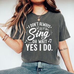 Pode incluir: Uma t-shirt de manga curta em cinzento-carvão com texto branco que diz I DON'T ALWAYS Sing ...OH WAIT YES I DO. O design inclui pautas e notas musicais acima da palavra Sing, com notas musicais adicionais nas laterais do texto.