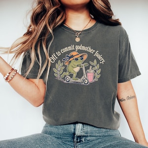 Könnte beinhalten: Ein anthrazitfarbenes Kurzarm-T-Shirt mit einer Grafik eines grünen Frosches, der einen Sonnenhut und eine Sonnenbrille trägt und auf einem Roller fährt. Der Text "Off to commit godmother foolery" steht darüber, daneben ein rosa Becher.
