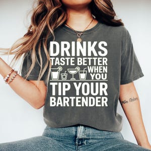 Könnte beinhalten: Dunkelgraues T-Shirt mit dem weißen Text "DRINKS TASTE BETTER WHEN YOU TIP YOUR BARTENDER". Das Shirt zeigt Cocktail-Illustrationen. Ein lässiges T-Shirt für Bar-Enthusiasten.