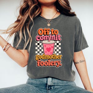 May include: A charcoal grey t-shirt featuring a graphic print of a pink iced coffee cup with hearts, set against a black and white checkered background. The text reads Off to com/mit godmother foolery in orange and pink retro-style lettering.