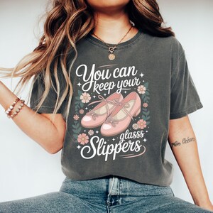 May include: Dark grey t-shirt featuring the text "You can keep your glasss Slippers" and a graphic of pink shoes. The design includes pink flowers and green leaves. The shirt is a casual style.