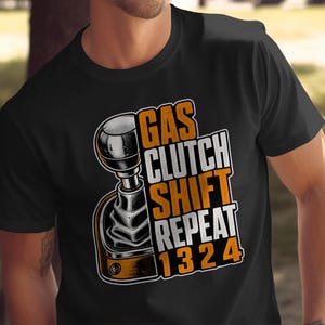 Könnte beinhalten: Ein schwarzes T-Shirt mit kurzen Ärmeln und einer Grafik eines manuellen Schalthebels. Das Design enthält den Text "GAS CLUTCH SHIFT REPEAT" in orangefarbenen und weißen Blockbuchstaben sowie die Zahlenfolge "1 3 2 4" in Orange darunter.