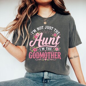 Könnte beinhalten: Ein anthrazitfarbenes Kurzarm-T-Shirt mit dem weißen und rosafarbenen Text I'M NOT JUST THE AUNT I'M THE GODMOTHER. Das Design ist mit rosa Rosen, grünen Blättern und kleinen Herzen verziert und mittig auf der Brust platziert.
