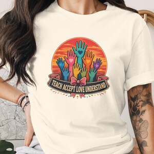 Könnte beinhalten: Ein cremefarbenes Kurzarm-T-Shirt mit einer kreisförmigen Grafik aus bunten erhobenen Händen mit kleinen Herzen. Unter den Händen zeigt ein Banner den Text TEACH ACCEPT LOVE UNDERSTAND in fetten Großbuchstaben mit einer rosa Schleife.