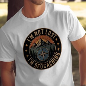 Puede incluir: Una camiseta blanca de manga corta con un gráfico circular en el pecho. El diseño muestra una cordillera, una brújula y el texto I'M NOT LOST I'M GEOCACHING en letra negrita, rodeado de pequeñas estrellas dentro de un borde negro.