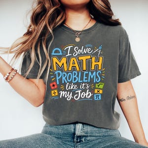 May include: A charcoal grey short-sleeve t-shirt featuring the text I Solve MATH PROBLEMS like it's my Job in yellow, blue, and white. The design includes colourful maths-themed icons such as a protractor, compass, square root, and pi symbol.