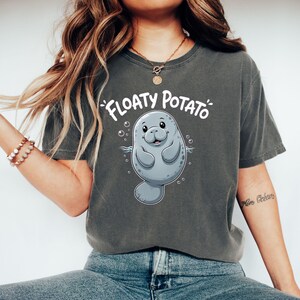 May include: A charcoal grey short-sleeve t-shirt featuring a graphic illustration of a light grey manatee surrounded by small bubbles. Above the manatee, the text "FLOATY POTATO" is printed in a bold, white, rounded, hand-lettered font style.
