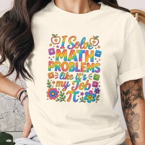 May include: A cream-coloured short-sleeve t-shirt featuring a colourful tie-dye graphic print that reads I Solve MATH PROBLEMS like it's my Job with various math symbols, apples, and flowers surrounding the text in a vibrant, multi-coloured design.