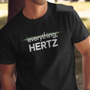 以下が含まれることがあります： 「everything, HERTZ」の白文字と緑色の波形グラフィックがプリントされた黒いTシャツ。半袖で、柔らかい素材で作られているように見える。
