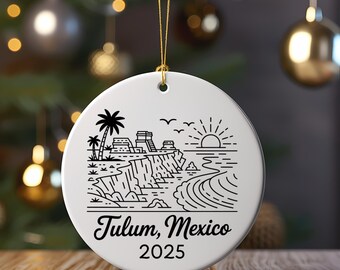May include: White ceramic Christmas ornament with a black line drawing of Tulum, Mexico, featuring palm trees, ancient ruins, and a sunrise over the ocean. The text "Tulum, Mexico 2025" is printed below the image. A gold string is attached for hanging.
