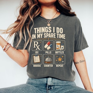 May include: A charcoal gray t-shirt featuring the text "THINGS I DO IN MY SPARE TIME" above a grid of six icons with labels: "RX" with a prescription symbol, "PILLS" with capsules, "BOTTLES" with medicine containers, "ORDERS" with a clipboard, "COUNTER" with a service bell, and "REPEAT" with a star.