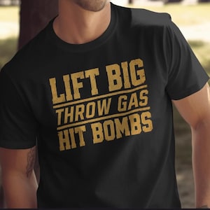 May include: A black short-sleeved t-shirt featuring the gold-colored text LIFT BIG THROW GAS HIT BOMBS printed in a distressed, block-letter style across the chest. The garment has a classic crew neckline and a standard fit.