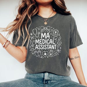 Puede incluir: Una camiseta de manga corta gris carbón con un gráfico circular blanco con el texto MA MEDICAL ASSISTANT rodeado de ilustraciones de un estetoscopio, un lazo, un portapapeles, una jeringa, un termómetro, una venda, guantes, un corazón y un frasco de medicina.