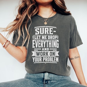 Peut inclure: Un t-shirt gris anthracite à manches courtes avec un texte blanc vieilli qui indique : SURE- LET ME DROP EVERYTHING AND WORK ON YOUR PROBLEM. Le t-shirt présente une encolure ronde et une coupe décontractée avec un motif graphique sur le devant.
