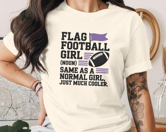 Puede incluir: Camiseta color crema con el texto "FLAG FOOTBALL GIRL (NOUN) SAME AS A NORMAL GIRL, JUST MUCH COOLER." El diseño incluye una bandera morada, un balón de fútbol y líneas horizontales moradas.