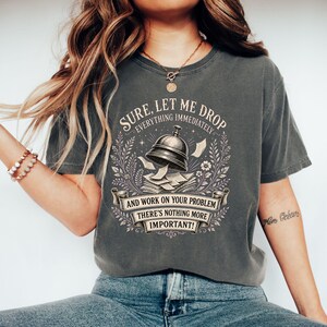 Könnte beinhalten: Ein anthrazitfarbenes T-Shirt mit kurzen Ärmeln und einer Grafik, die eine Tischklingel und verstreute Papiere zeigt. Der Text lautet: SURE, LET ME DROP EVERYTHING IMMEDIATELY AND WORK ON YOUR PROBLEM THERE'S NOTHING MORE IMPORTANT!