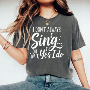 Könnte beinhalten: Ein anthrazitfarbenes Kurzarm-T-Shirt mit weißem Text, der I DON'T ALWAYS Sing... OH WAIT, Yes I Do lautet, begleitet von kleinen Notengrafiken. Das T-Shirt hat einen Rundhalsausschnitt und eine lockere Passform aus Baumwollstoff.