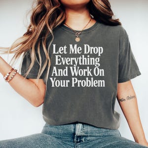 Könnte beinhalten: Ein anthrazitfarbenes Kurzarm-T-Shirt mit dem weißen Text Let Me Drop Everything And Work On Your Problem in Serifenschrift auf der Brust. Das T-Shirt hat einen Rundhalsausschnitt und eine lockere Passform, kombiniert mit einer blauen Jeans.