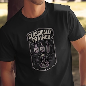 Może przedstawiać: Czarny t-shirt z krótkim rękawem z białym nadrukiem graficznym przedstawiającym manualną dźwignię zmiany biegów i trzy pedały. Napis "CLASSICALLY TRAINED" jest wydrukowany pogrubioną czcionką nad techniczną ilustracją zamkniętą w obrysie przypominającym odznakę.
