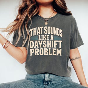 Könnte beinhalten: Ein anthrazitfarbenes Kurzarm-T-Shirt mit dem Text "THAT SOUNDS LIKE A DAYSHIFT PROBLEM" in cremefarbener Blockschrift, ergänzt durch zwei horizontale türkisfarbene Linien zwischen den Wörtern "LIKE A" und "DAYSHIFT".