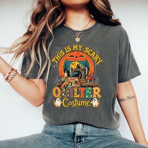 Könnte beinhalten: Ein anthrazitfarbenes T-Shirt mit einem Grafikdruck, der einen Kürbiskopf, eine Nähmaschine, einen bunten Patchwork-Quilt und den Text "THIS IS MY SCARY QUILTER Costume" sowie zwei kleine Geisterillustrationen am unteren Rand zeigt.
