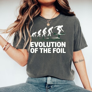 May include: Dark grey t-shirt with the text "EVOLUTION OF THE FOIL" in white. The graphic shows the evolution of a person foil surfing. The shirt is a comfortable, casual style.