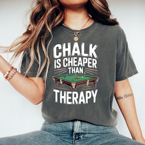 May include: Dark gray t-shirt with the text "CHALK IS CHEAPER THAN THERAPY" in white. The graphic includes a pool table with balls and cues. The shirt is a comfortable, casual style.